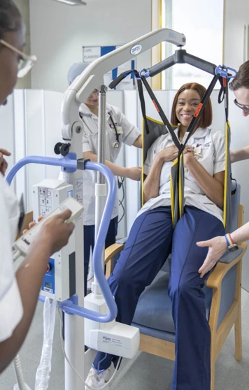 Care students practice using a patient hoist with a seated classmate in a simulated care setting. Care students practice using a patient hoist with a seated classmate in a simulated care setting.